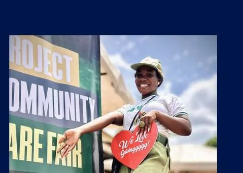 A corps member holding a heart-shaped sign beside a Community Care Fair banner