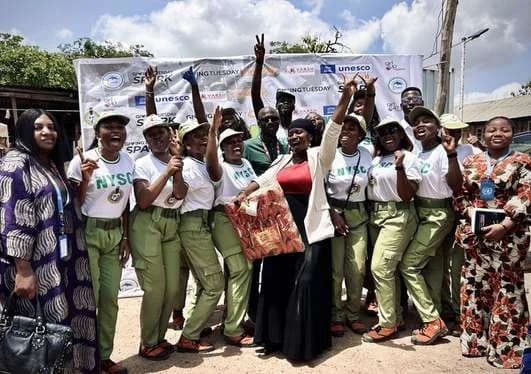 Corps members in NYSC uniform celebrating with community members in front of a banner
